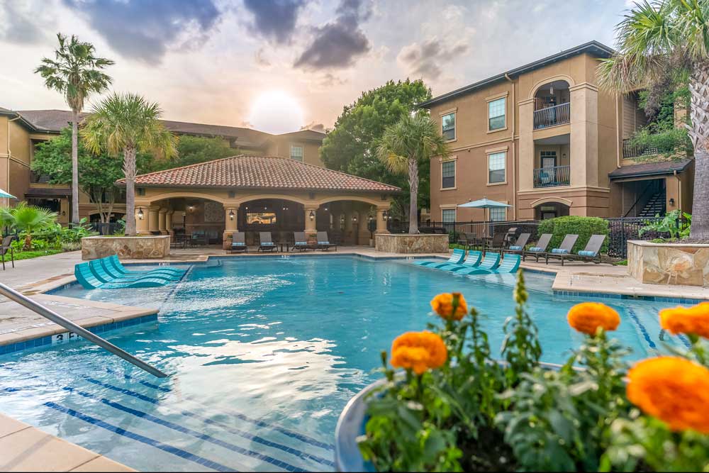 Swimming pool at sunset with in-water loungers, palm trees, and apartment buildings surrounding landscaped courtyard