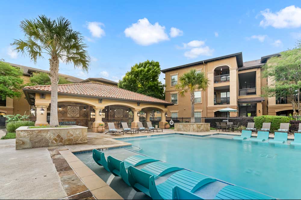 Daytime pool with in-water loungers, palm trees, and apartment buildings with balconies surrounding courtyard