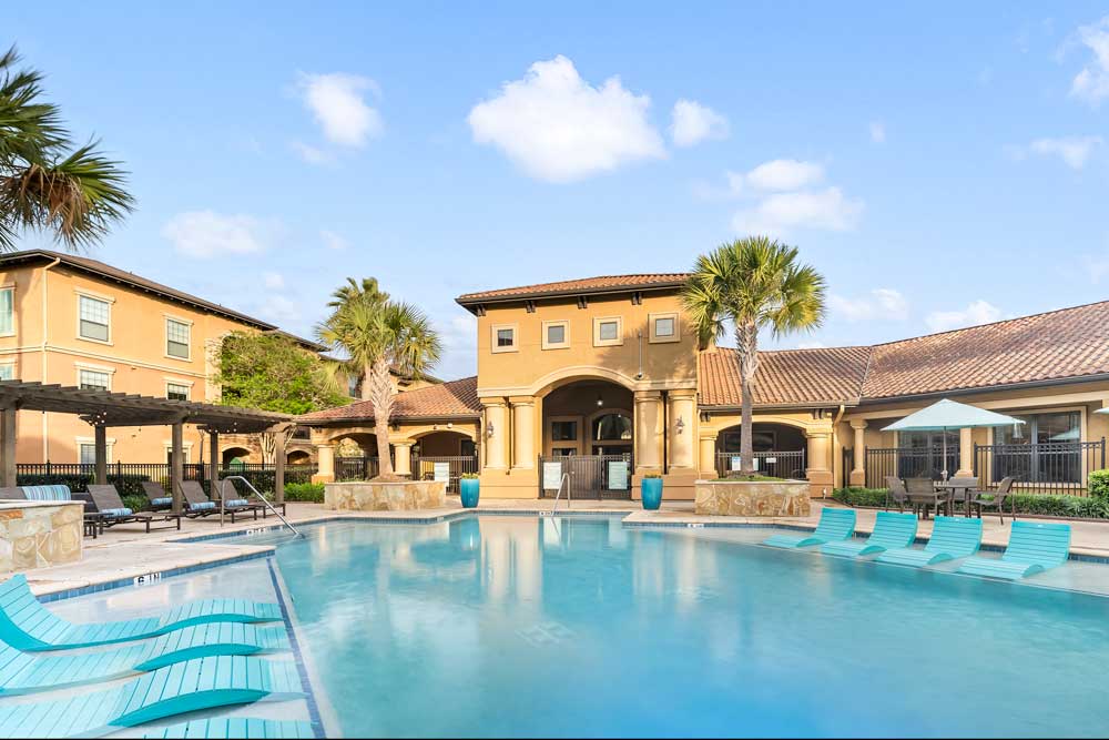 Pool courtyard with in-water loungers, clubhouse with arched entry, palm trees, and shaded seating areas with umbrellas