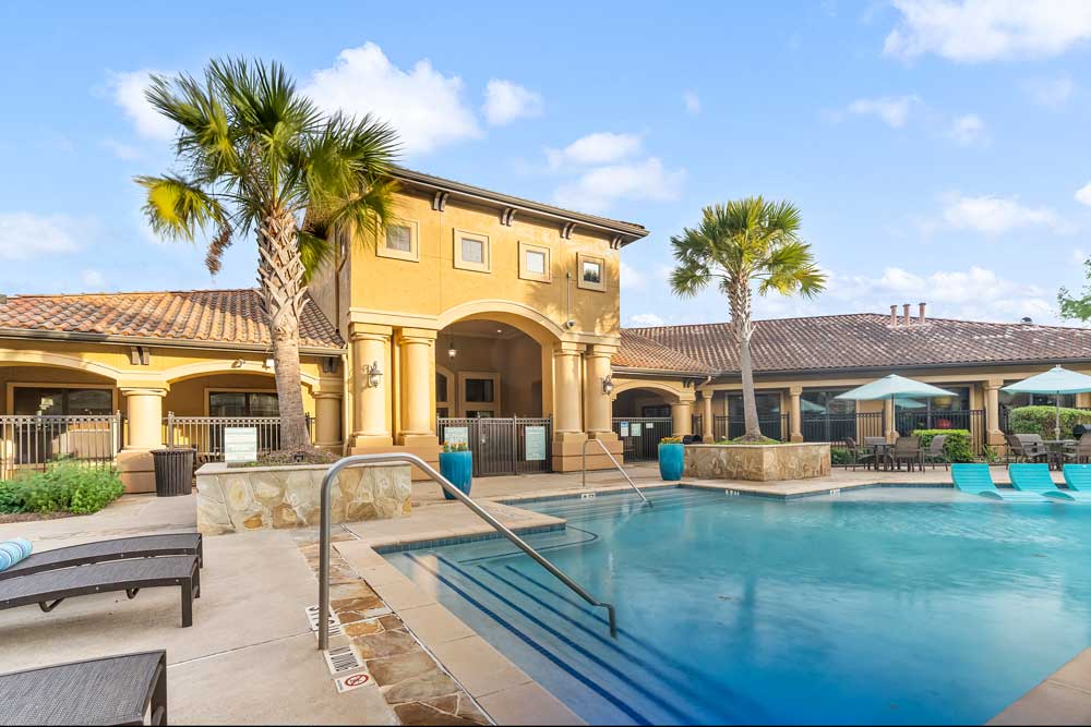 Pool with entry steps and handrails, clubhouse with arched columns, palm trees, and lounge seating along deck