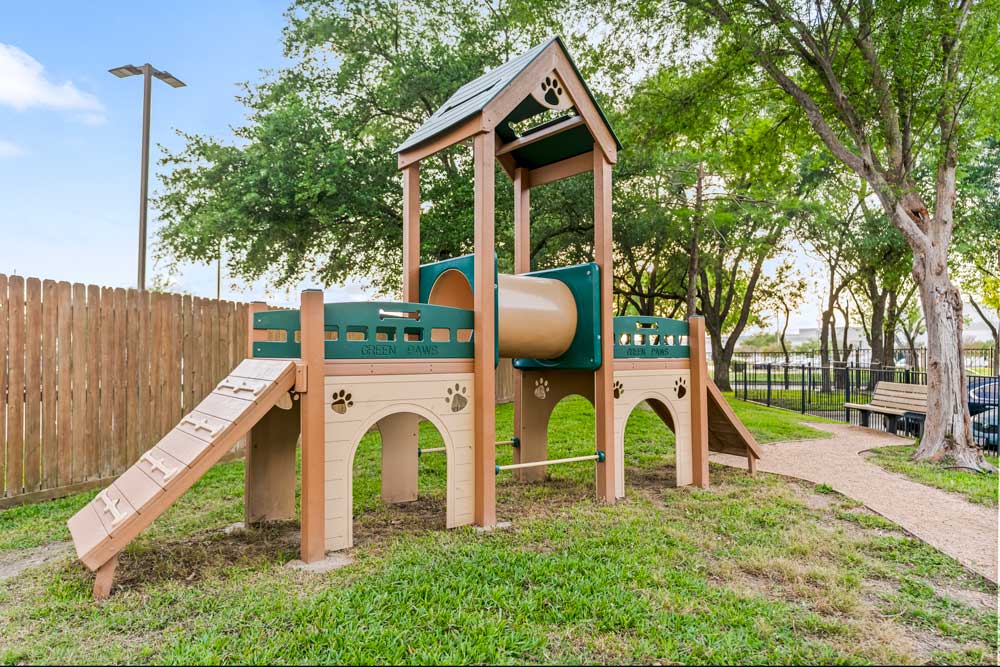 Pet play structure with tunnel and ramp, grassy area, wooden fence, and walking path under tall trees