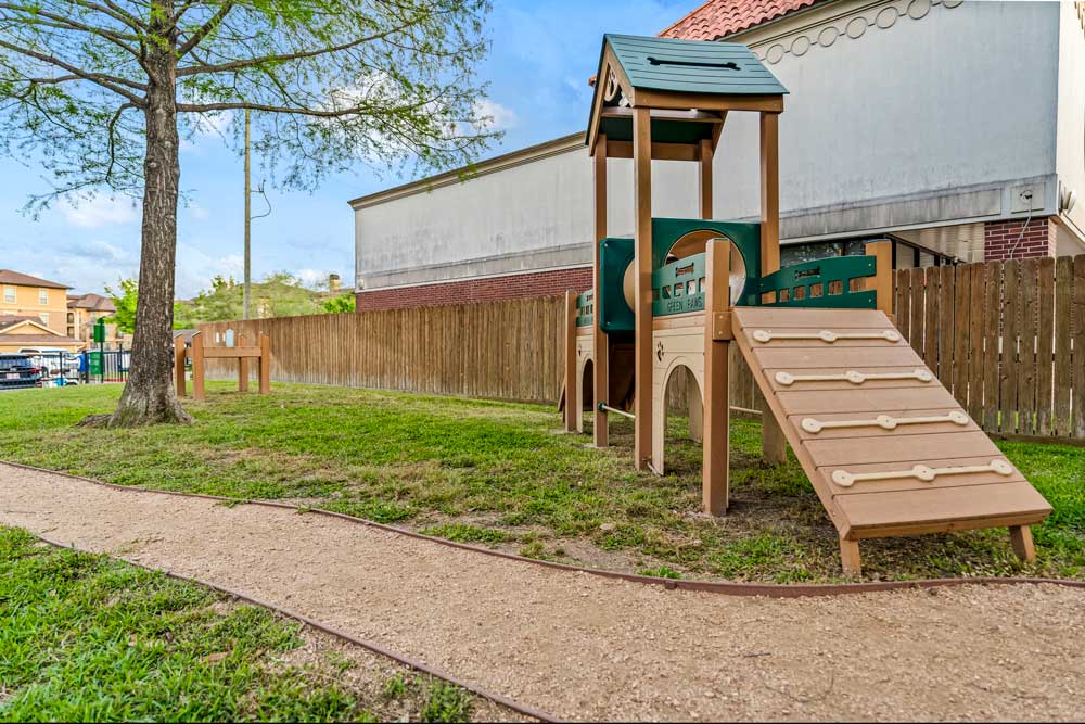 Dog park with ramp play structure, fenced perimeter, and gravel path winding through shaded green space