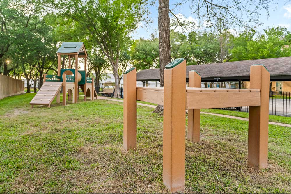 Dog park with agility platform, play structure, and shaded grassy area enclosed by fencing and mature trees