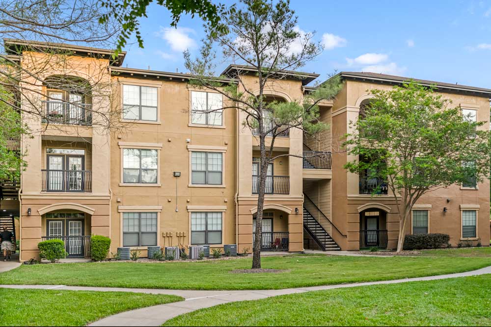 Apartment community exterior with multiple buildings, arched balconies, green lawn areas, and mature trees throughout property