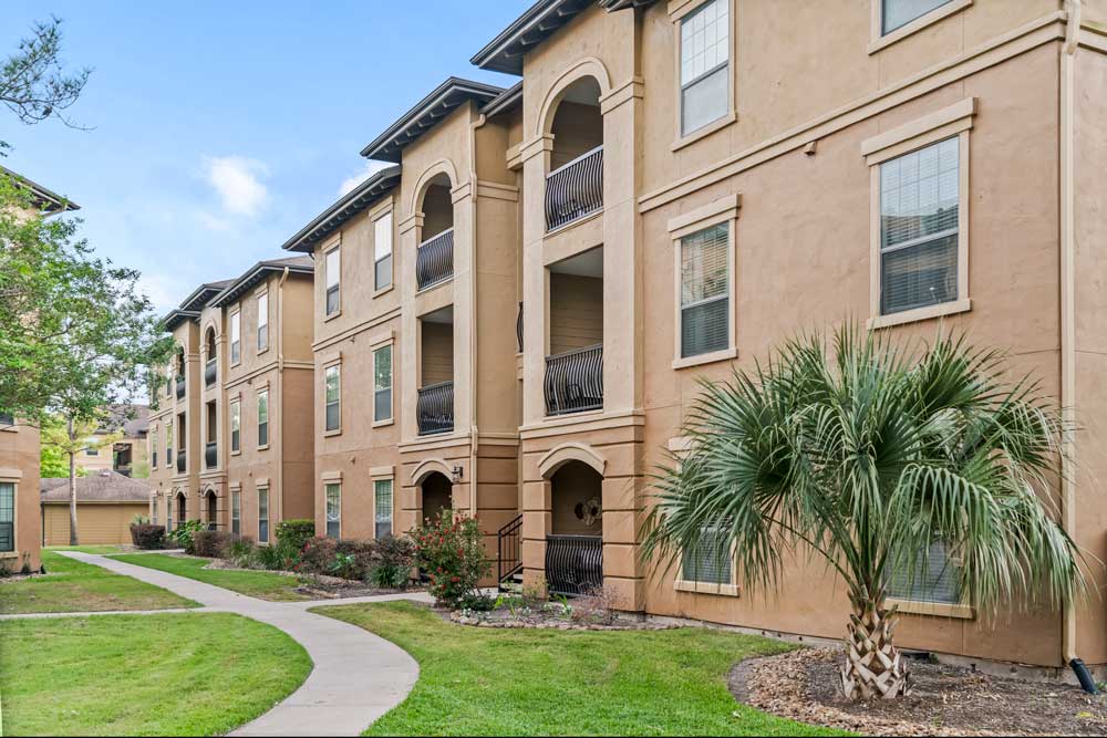 Three-story apartment building with stucco exterior, arched balconies, landscaped lawn, and curved sidewalk pathway