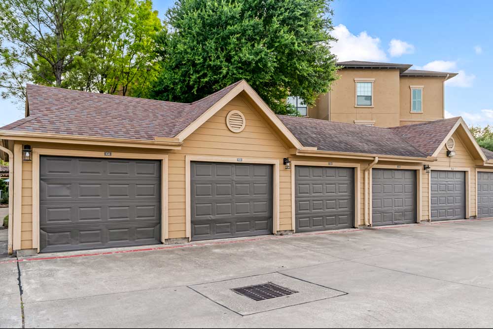 Row of detached garages with dark doors, pitched roofs, and concrete drive set against trees and apartment buildings