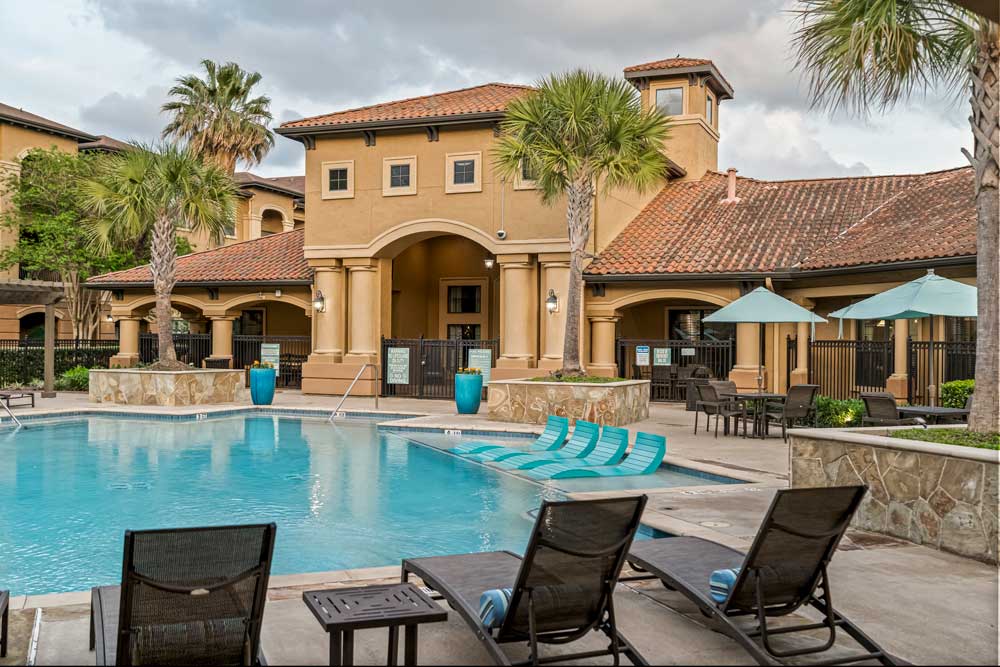 Resort-style pool with lounge chairs, umbrellas, palm trees, and clubhouse featuring arched entry and tiled roof