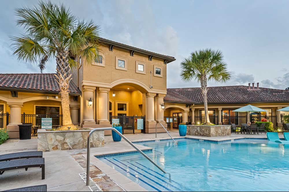 Apartment pool with steps, handrails, palm trees, and clubhouse with covered entry and outdoor seating areas