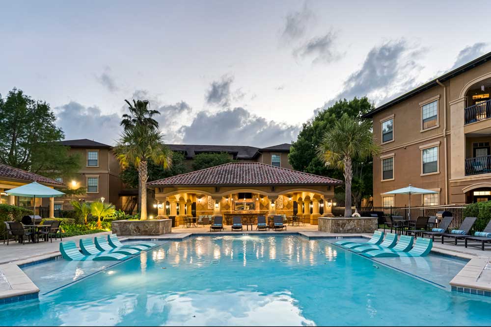 Symmetrical pool view with in-water loungers, palm trees, and warmly lit clubhouse centered within landscaped courtyard