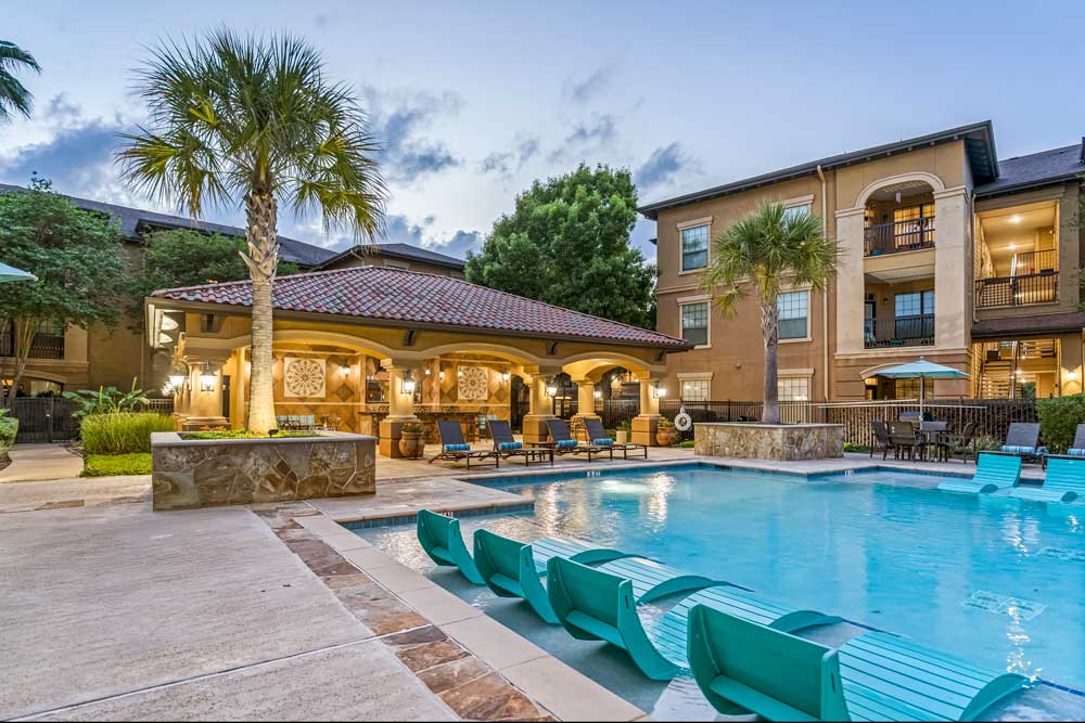 Pool area with in-water loungers, palm trees, and illuminated clubhouse with seating and umbrellas during early evening light
