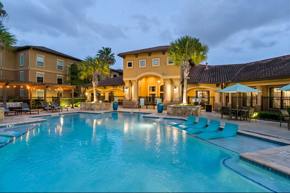 Apartment pool with in-water loungers, palm trees, and lit clubhouse with outdoor seating creating a calm evening setting