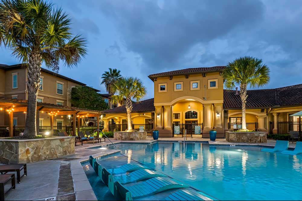 Swimming pool with in-water loungers, pergola seating area, and illuminated clubhouse creating a relaxed evening atmosphere
