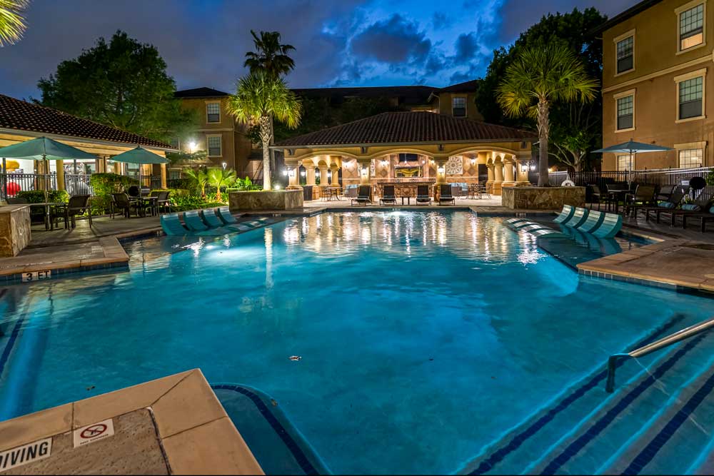 Resort-style pool with steps, in-water loungers, shaded seating, and clubhouse with warm lighting surrounded by palm trees