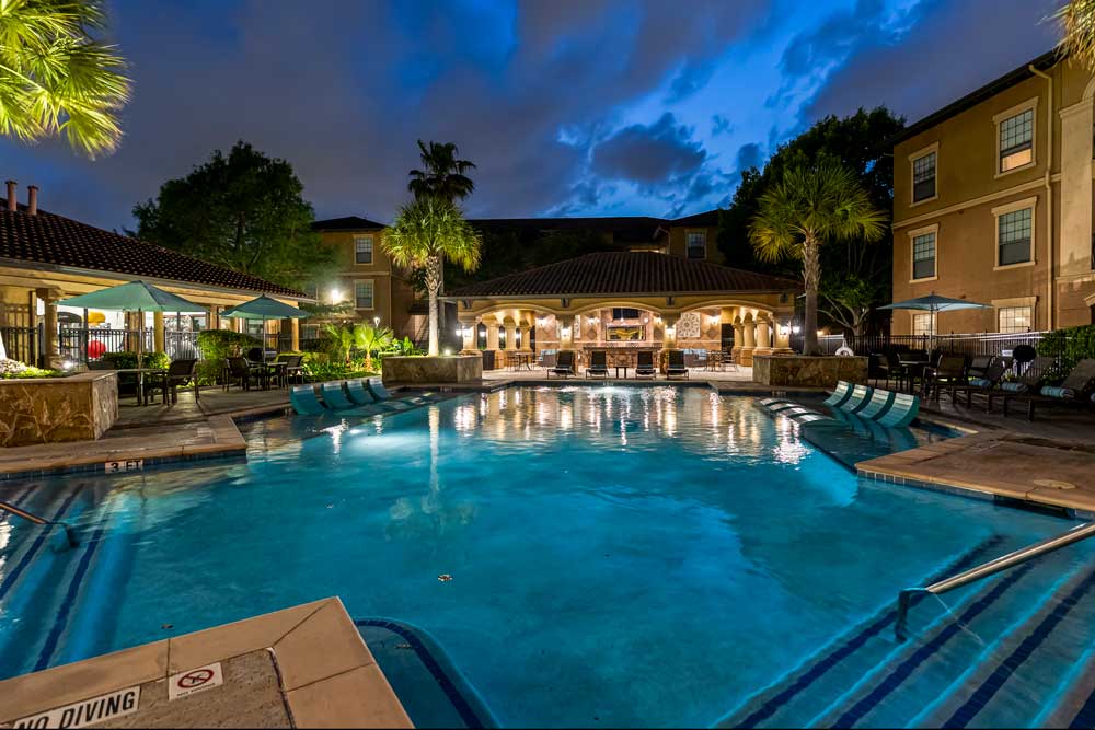 Resort-style pool with curved edges, in-water loungers, shaded seating, and lit clubhouse framed by palm trees at dusk