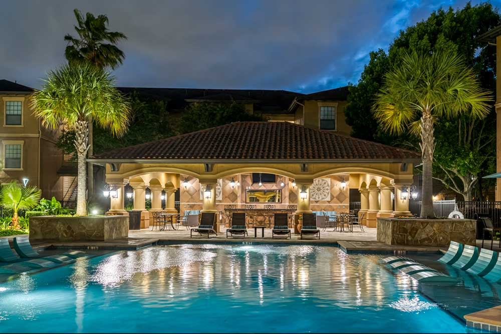 Poolside pavilion with bar seating, lounge chairs, and palm trees reflected in water, surrounded by apartment buildings at night