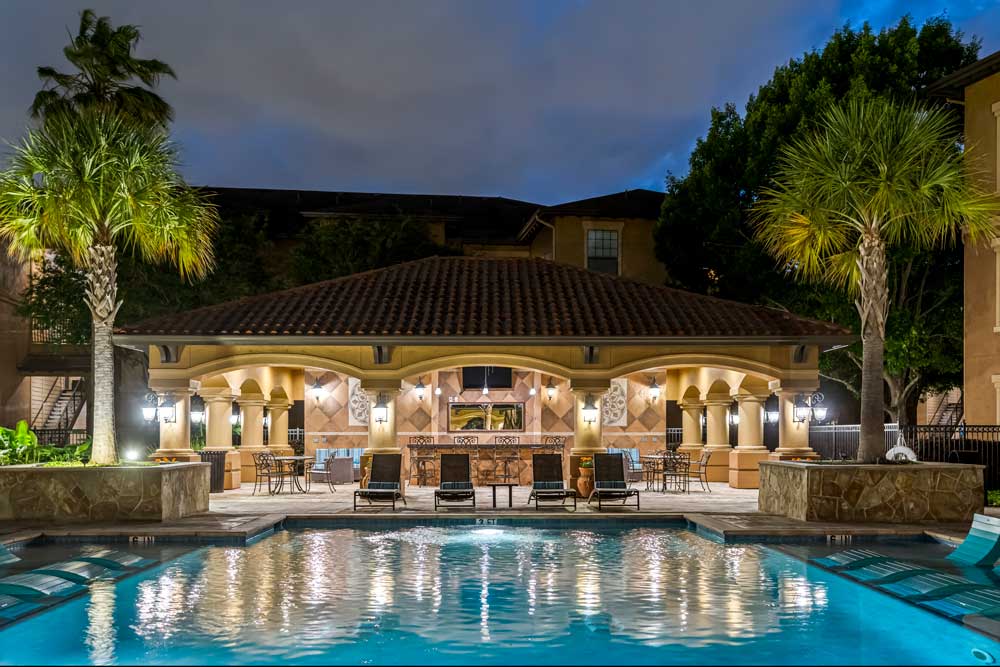 Poolside pavilion with seating, bar area, and lighting reflected on water, surrounded by palm trees and landscaped courtyard at night