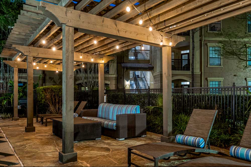 Pergola lounge with string lights, cushioned seating, and chaise chairs beside landscaped fencing and apartment buildings at night