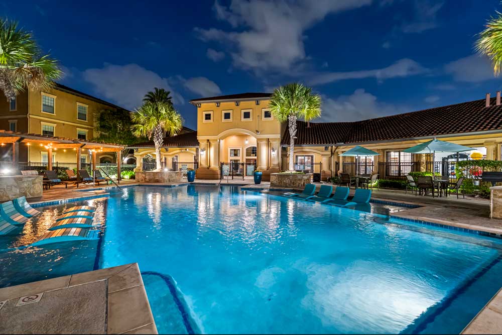 Expansive swimming pool with in-water loungers, shaded seating areas, and lit clubhouse surrounded by landscaped grounds at night