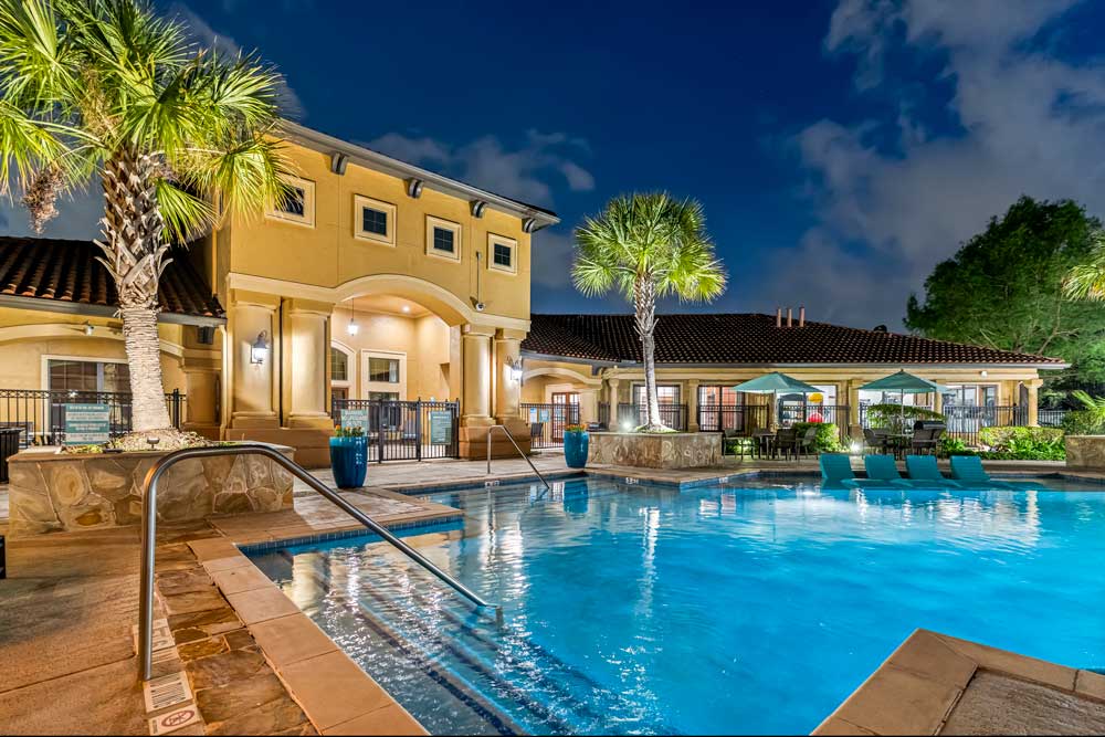 Resort-style pool with illuminated clubhouse, palm trees, lounge seating, and umbrellas creating a relaxing evening atmosphere