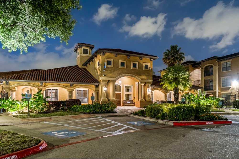 Illuminated community clubhouse with covered entry, landscaped greenery, palm trees, and accessible parking spaces in front