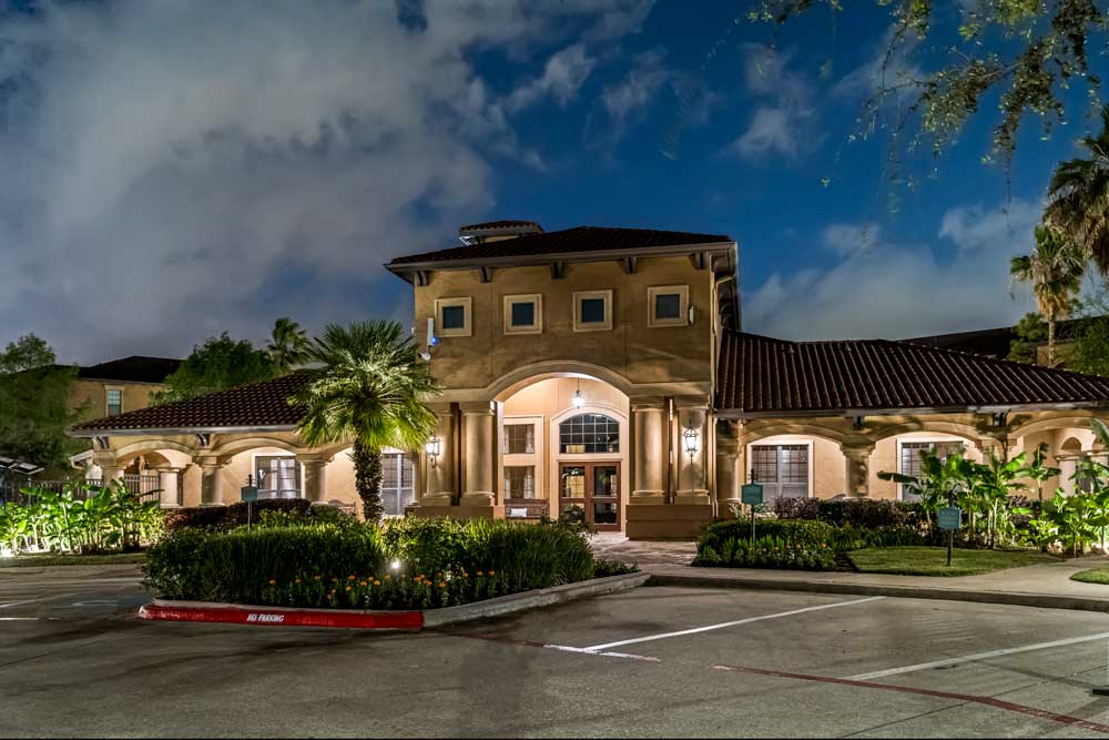Elegant clubhouse exterior at dusk with arched entryway, warm lighting, tiled roof, and landscaped grounds surrounding parking area