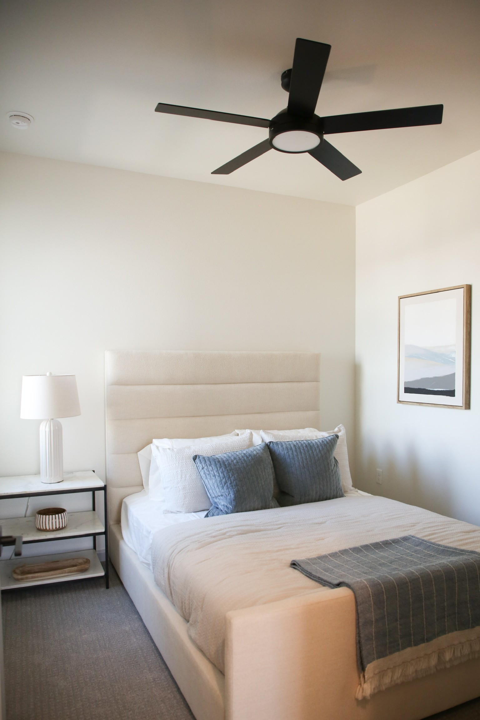 Bright and airy bedroom with upholstered headboard, blue accent pillows, ceiling fan, and stylish nightstand 