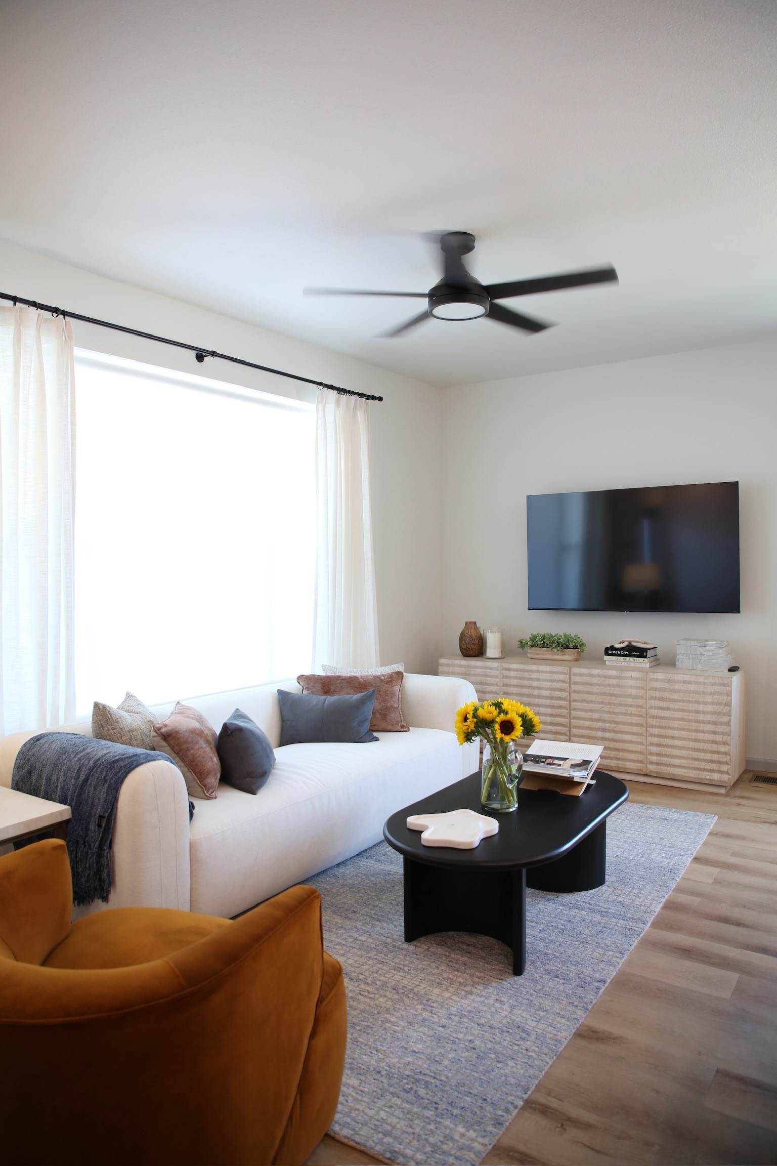 Open living room with white sofa, hardwood-style floors, ceiling fan, large window, and wall-mounted TV 