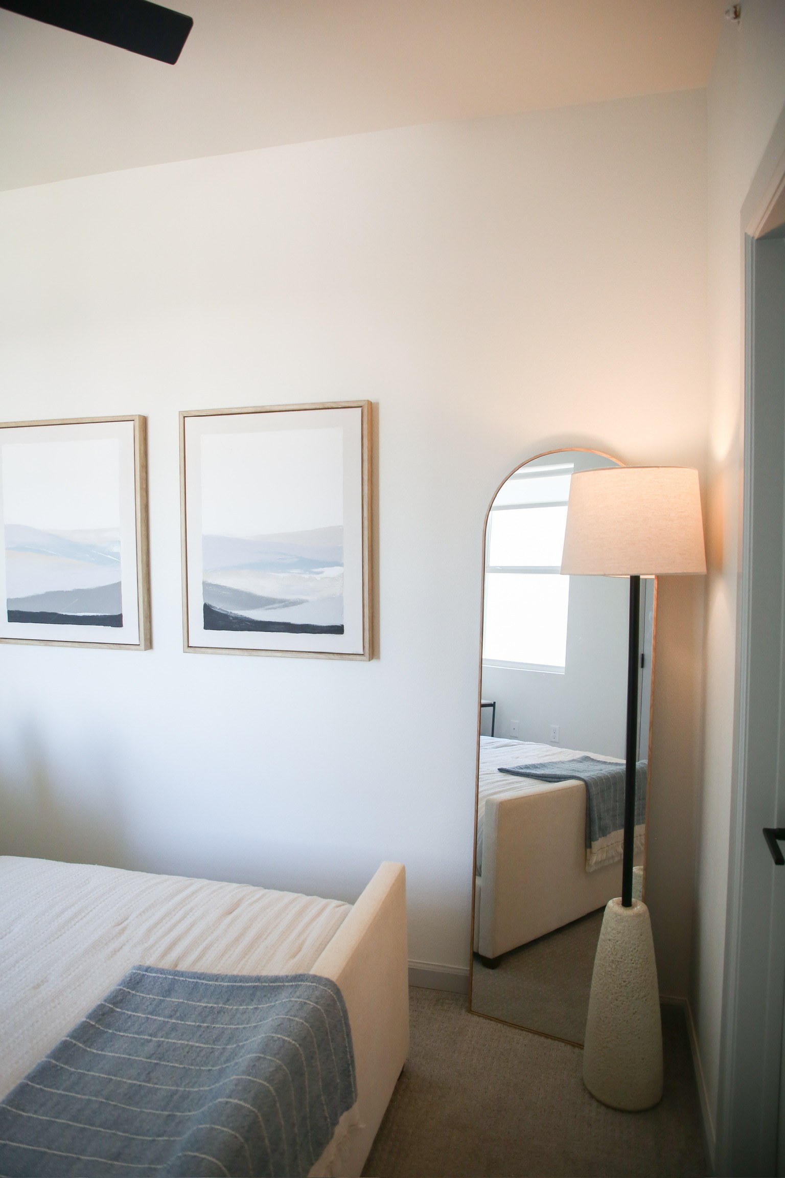 Bright bedroom corner with paired landscape artwork, arched floor mirror, and floor lamp alongside an upholstered bed
