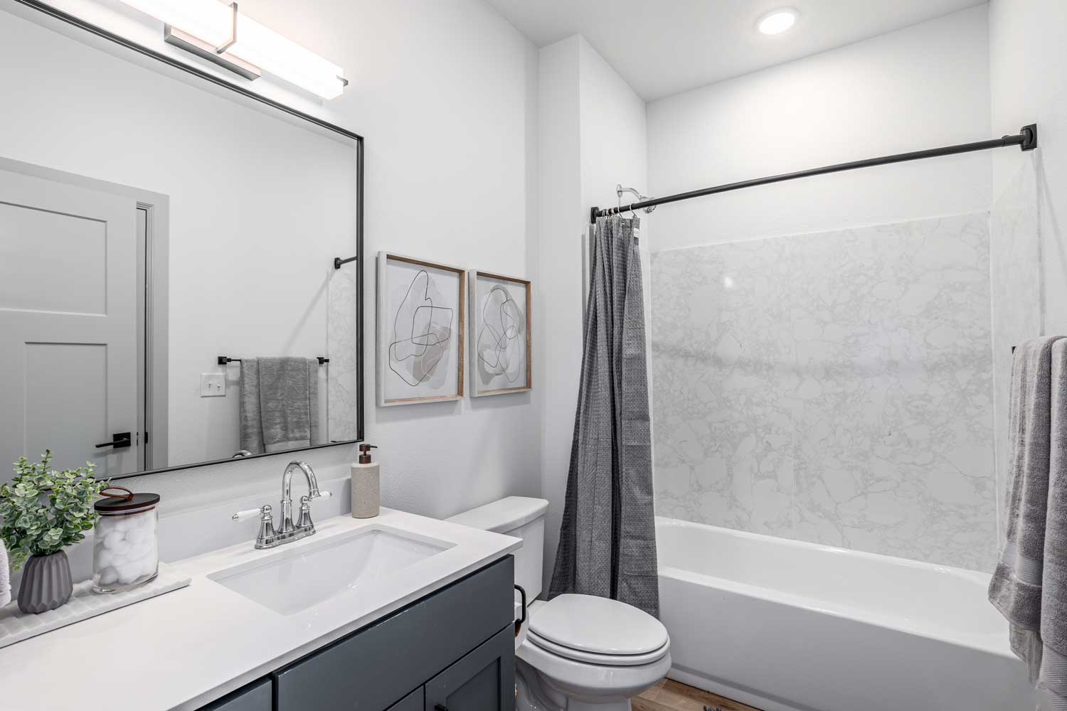 Bright bathroom with oversized mirror, gray vanity cabinet, marble-look tub surround, and decorative wall art