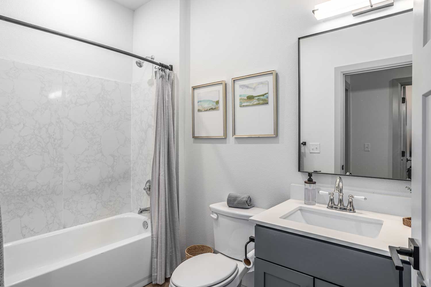 Bright bathroom with gray vanity cabinet, framed artwork, marble-look tub surround, and modern black fixtures
