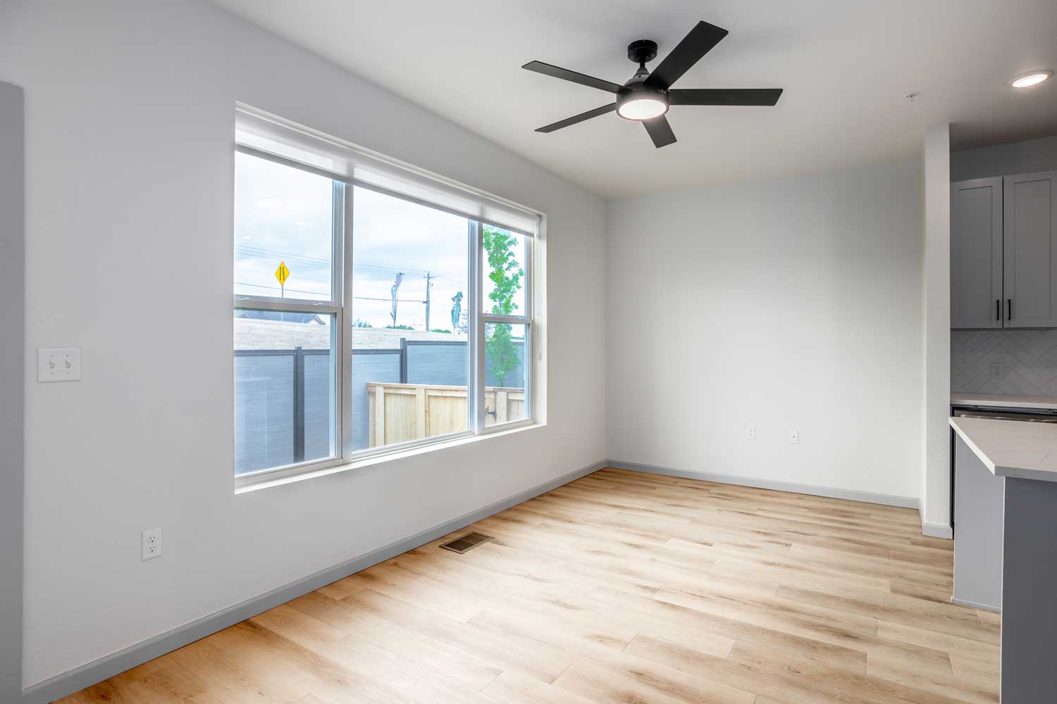 Open living area with wood-style flooring, ceiling fan, oversized window, fenced outdoor space, and neutral walls
