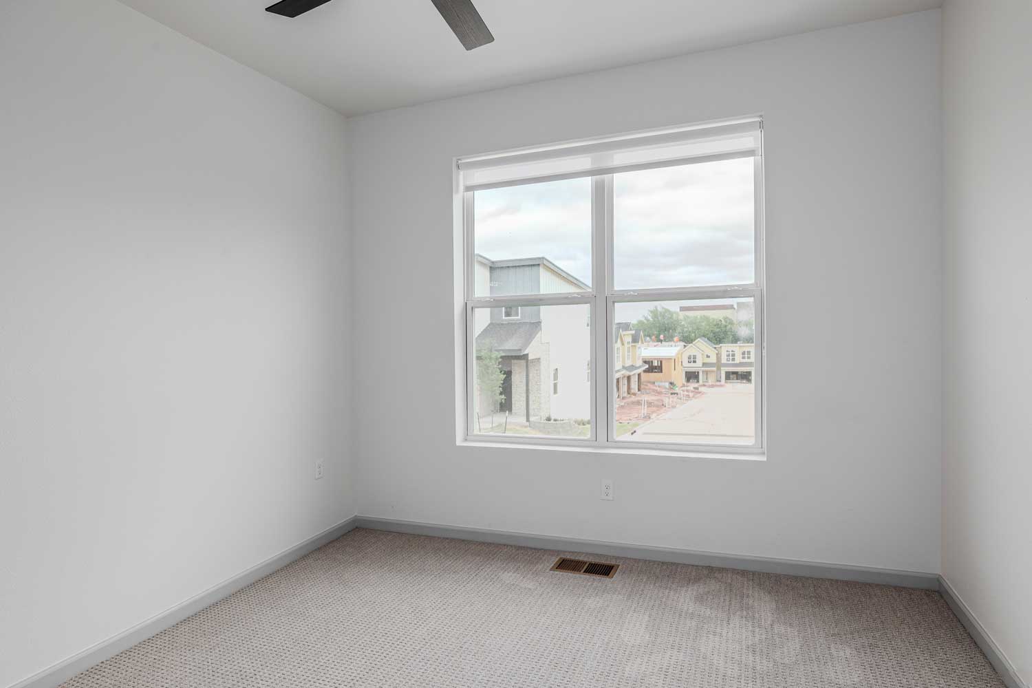 Bright bedroom with soft carpet flooring, large window, neutral painted walls, and view of nearby residential buildings