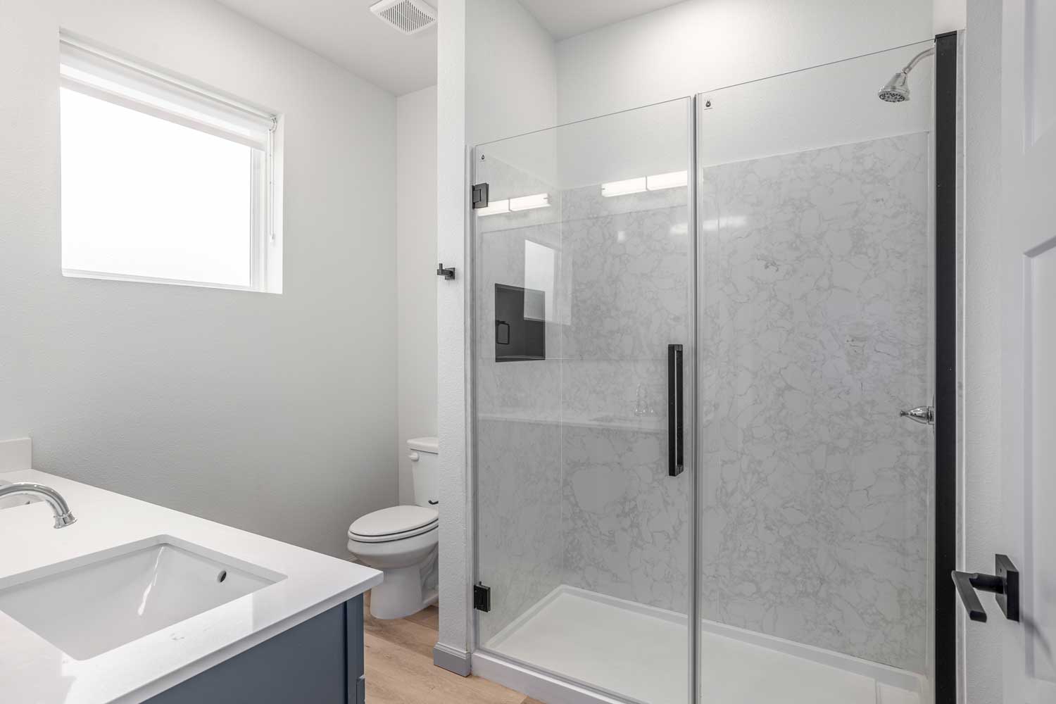 Modern bathroom with glass shower enclosure, marble-look wall panels, white vanity counter, and frosted window