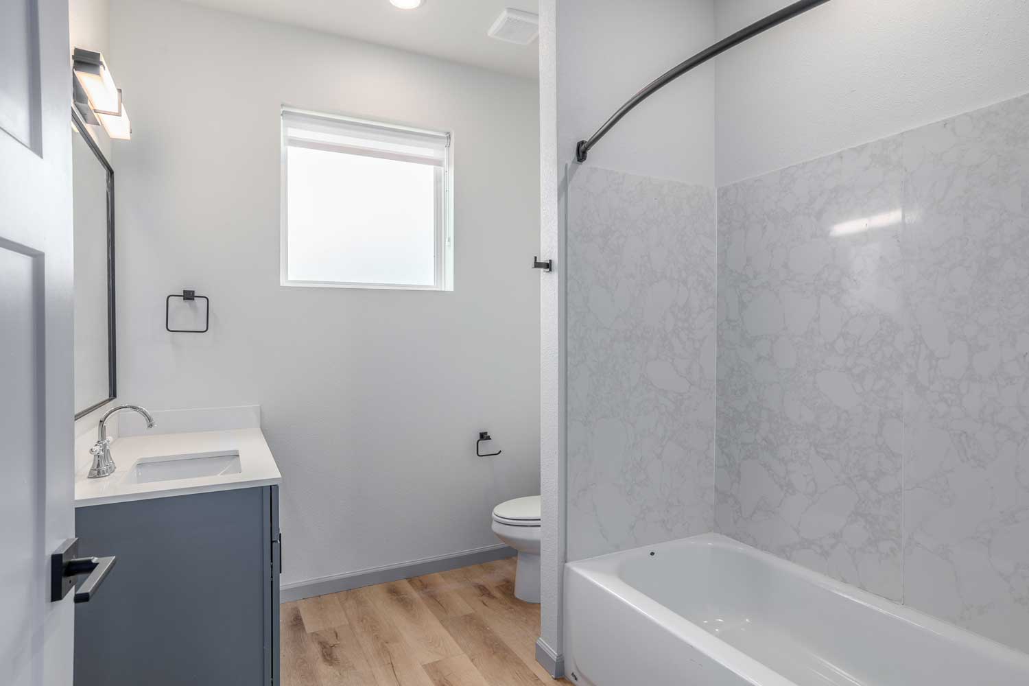 Bright bathroom with marble-look tub surround, gray vanity cabinet, frosted window, and wood-style flooring below