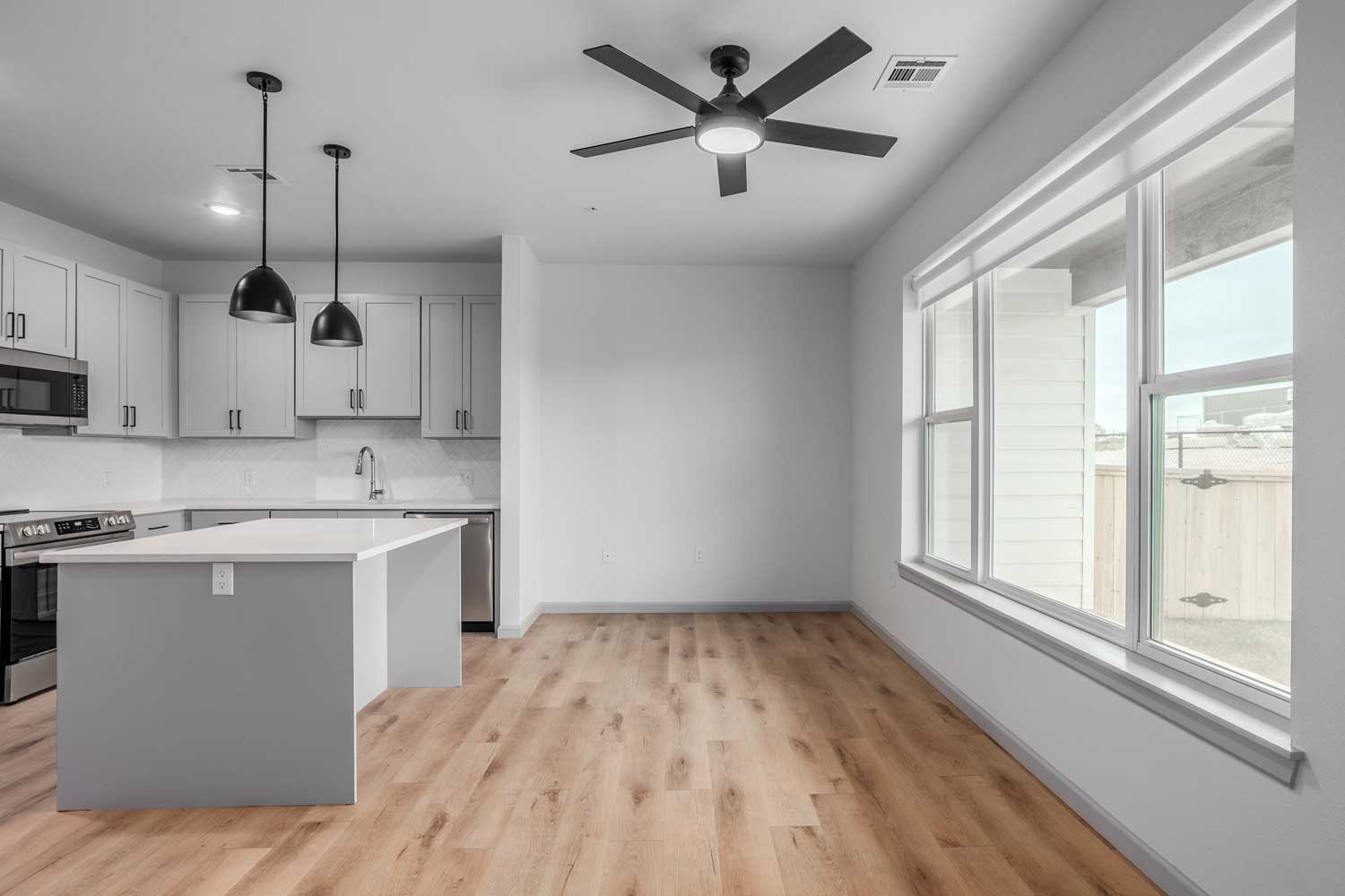 Open living area with kitchen island, pendant lighting, wide windows, wood-style flooring, and ceiling fan overhead above