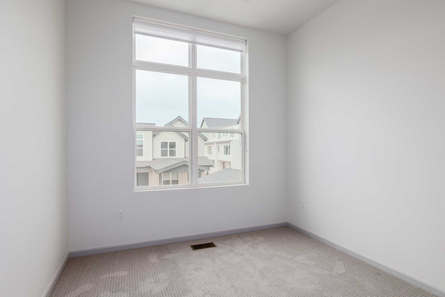 Minimal bedroom with carpet flooring, tall window, neutral walls, ceiling fan, and view of neighboring homes outside nearby