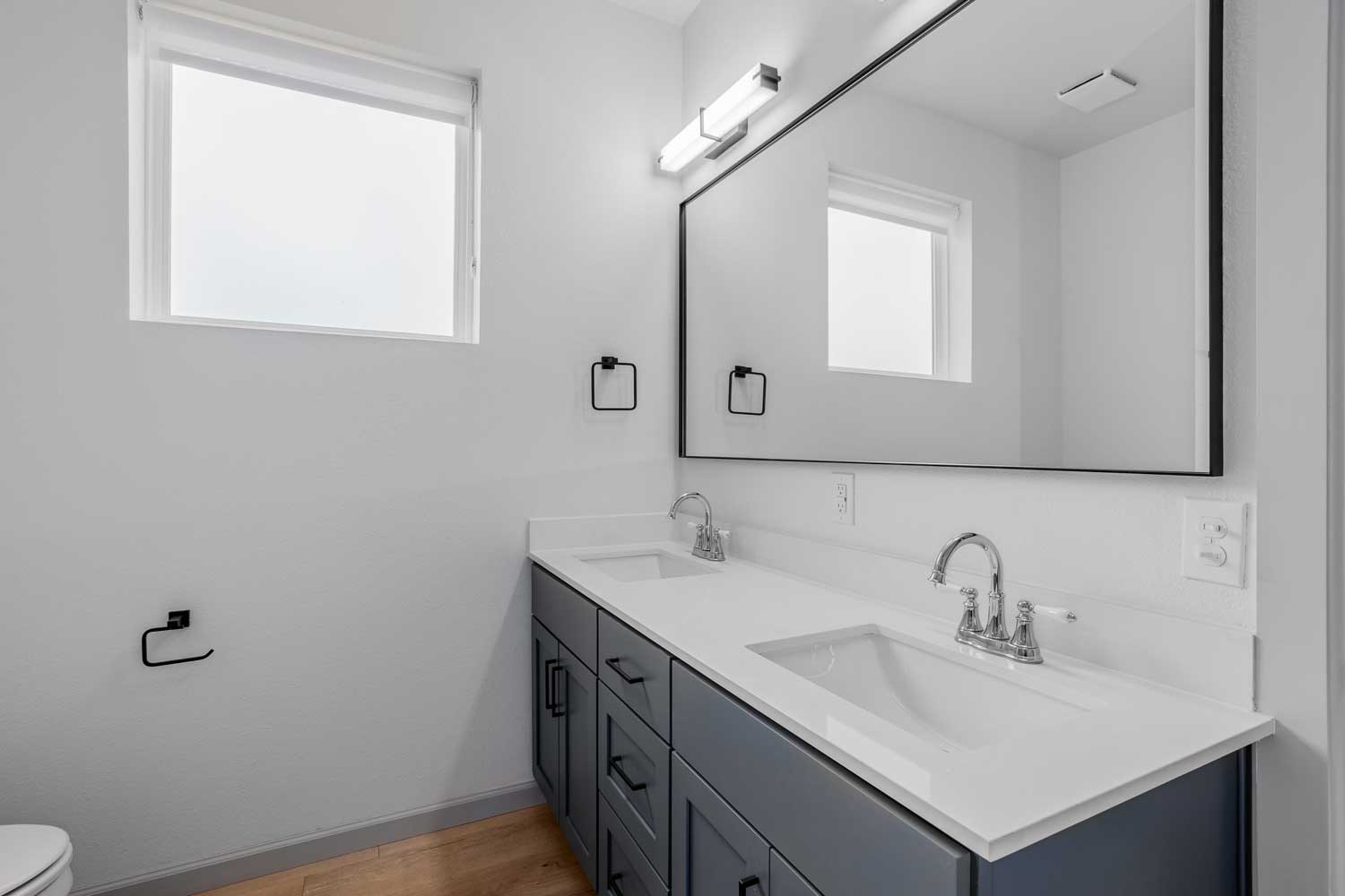 Modern bathroom with double sink vanity, large framed mirror, black fixtures, frosted window, and wood-style flooring below