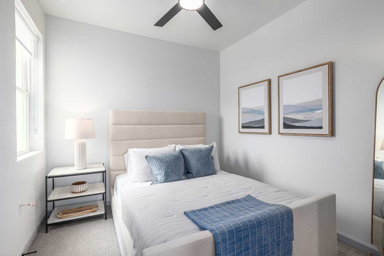 Bright bedroom with upholstered bed, blue accent pillows, framed wall art, side table, tall window, and soft carpeting throughout