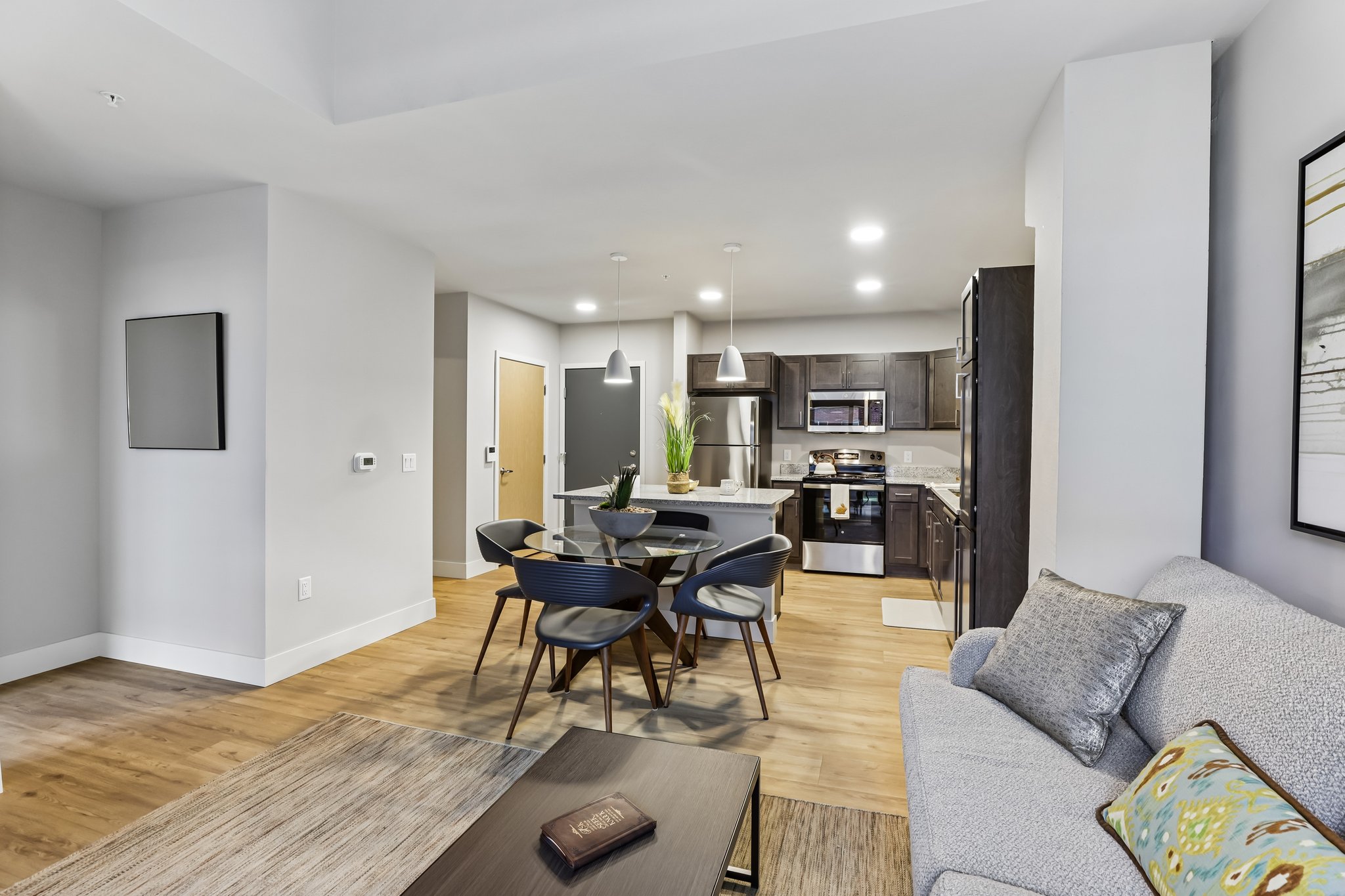 Living room into Kitchen in a one-bedroom income-restricted 55+ senior apartment at The Victoria at Huxley Yards in Madison, WI, with open layout, carpeted flooring, and natural light.
