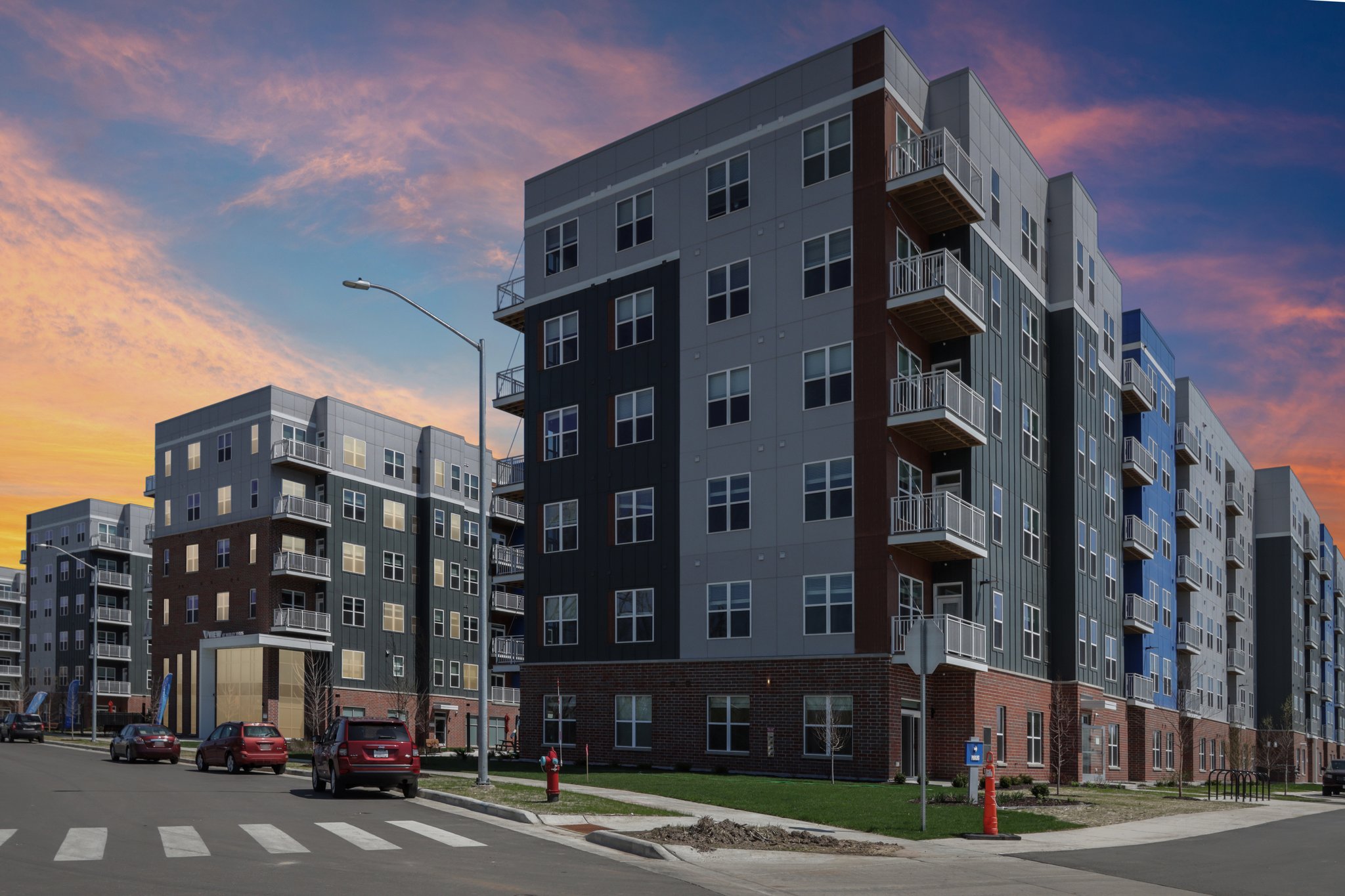 Street-level view of modern apartment buildings with colorful facades, landscaped walkways, residents biking and walking, and a pool courtyard