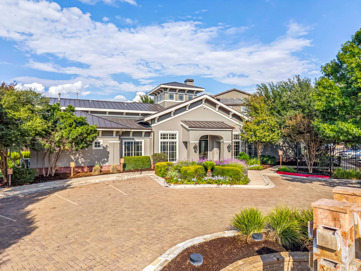 Front view of a gray stucco apartment leasing office with a metal roof, brick paver lot, and manicured landscaping