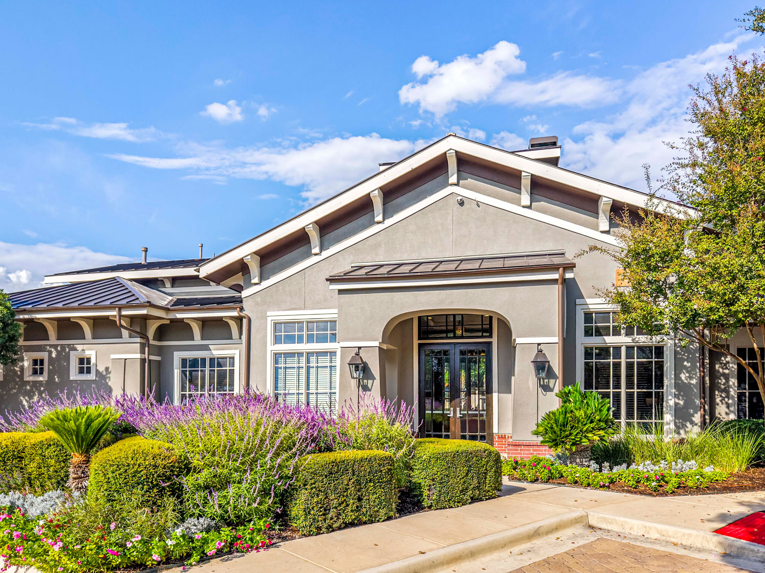 Daytime exterior of a gray stucco leasing office building with an arched entrance, purple sage, and trimmed hedges