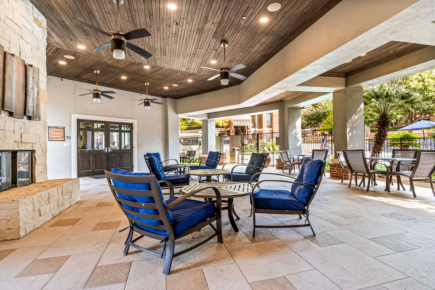 Covered patio lounge with blue cushioned seating, ceiling fans, stone fireplace, and outdoor dining tables in courtyard setting