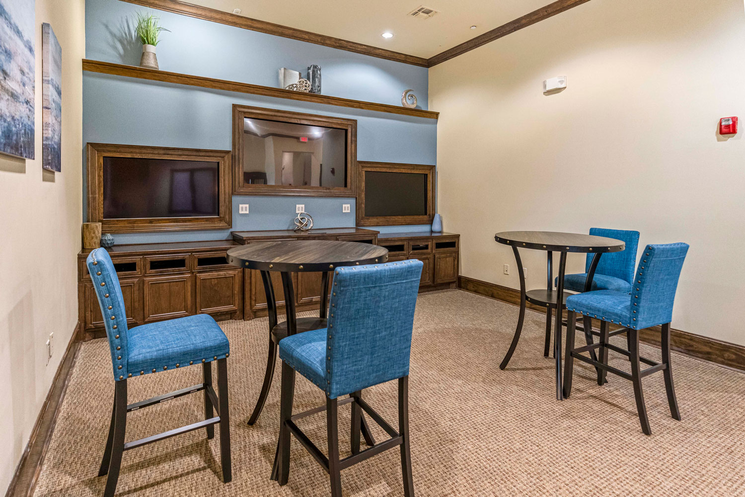 Community game room with high-top tables, blue upholstered chairs, built-in media wall, shelving decor, and soft carpet flooring