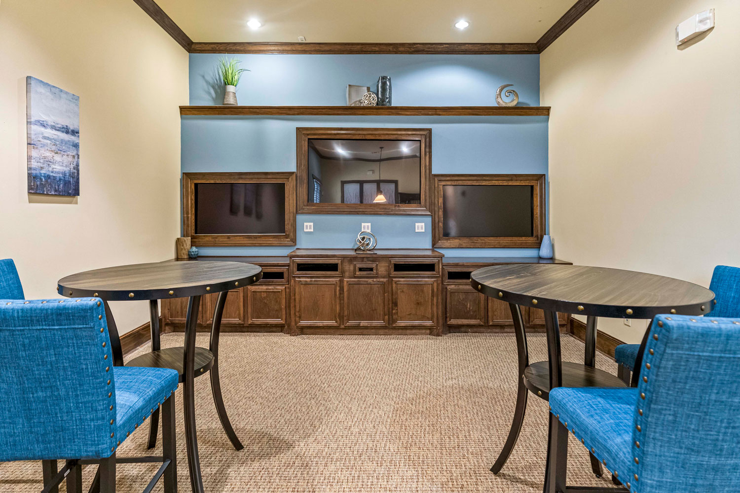 Resident lounge with round tables, blue upholstered chairs, built-in entertainment wall, and carpeted clubhouse interior