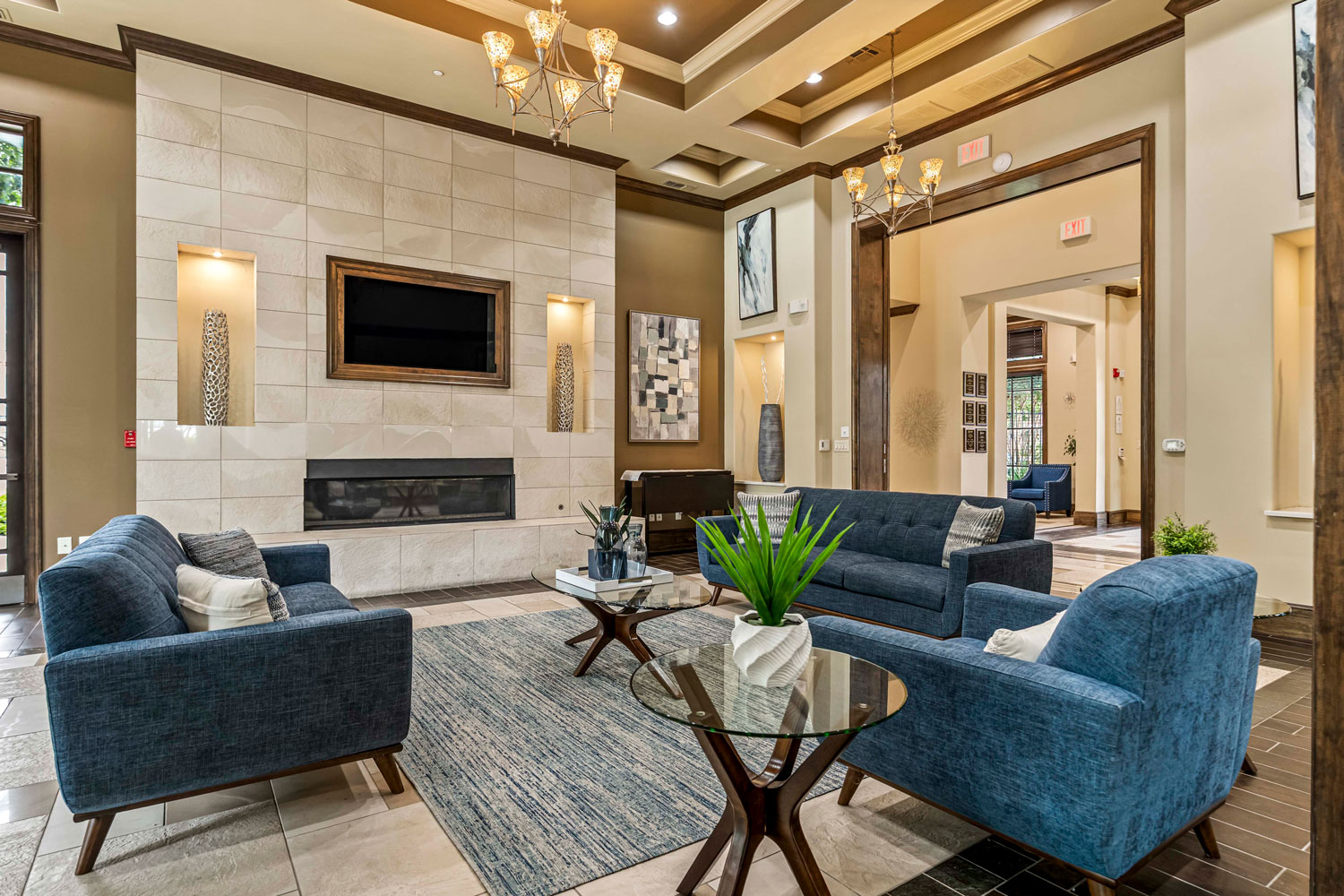 Spacious resident lounge with blue sofas, glass coffee tables, fireplace feature wall, decorative lighting, and open lobby access