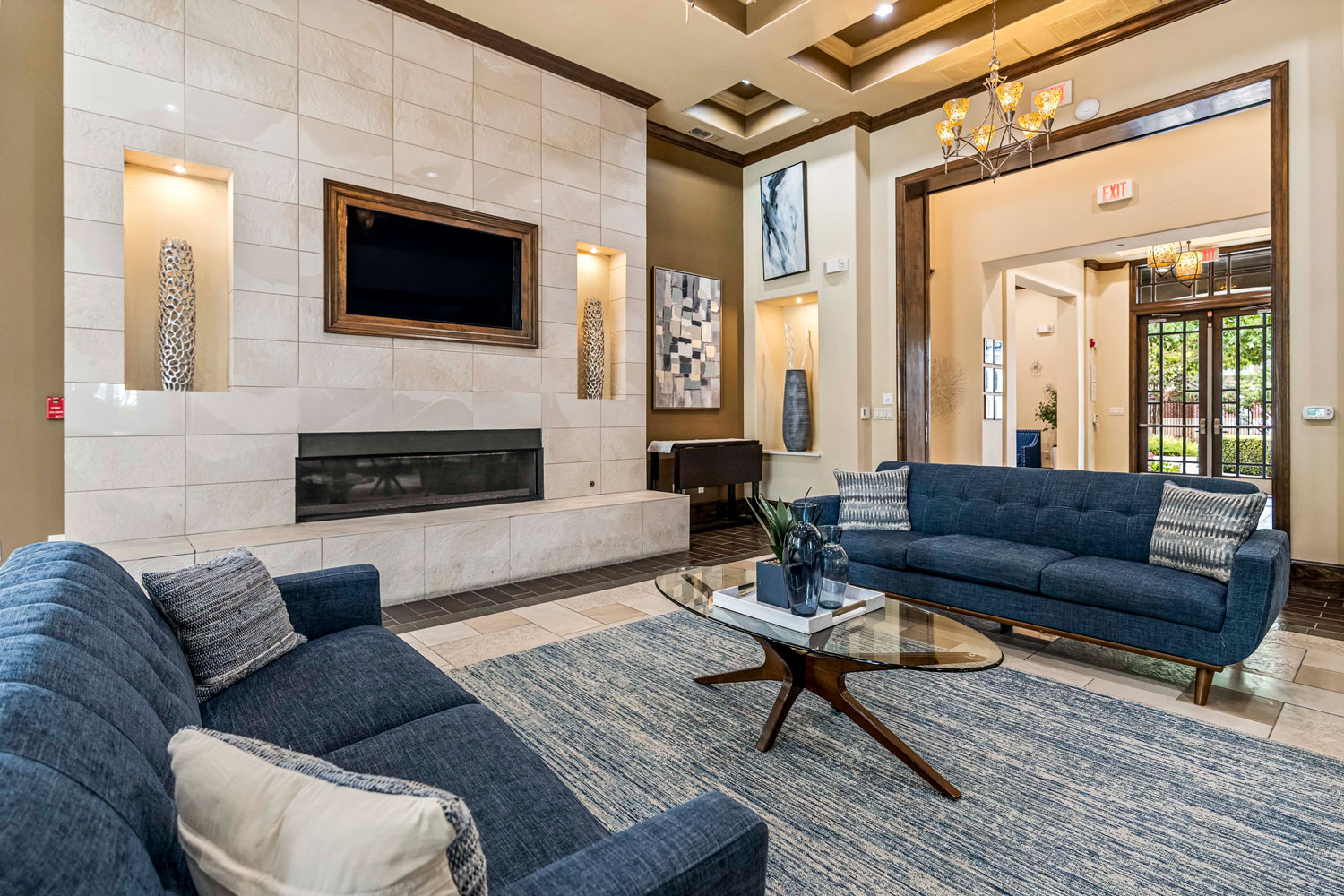 Clubhouse lounge with blue sofas, glass coffee table, fireplace feature wall, art accents, and open hallway leading to lobby entrance