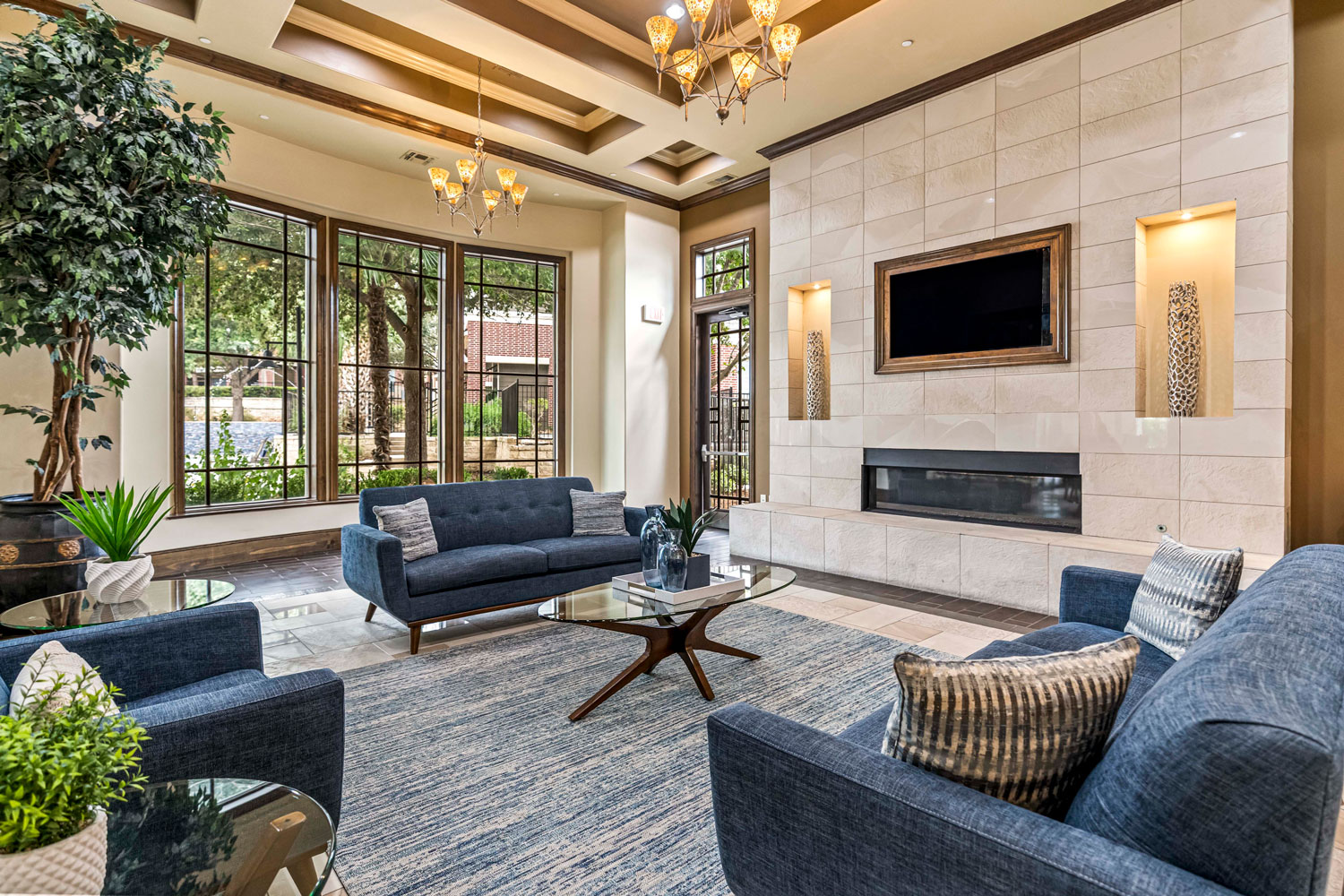 Resident clubhouse lounge with blue sofas, glass coffee tables, fireplace feature wall, coffered ceiling, and large courtyard windows