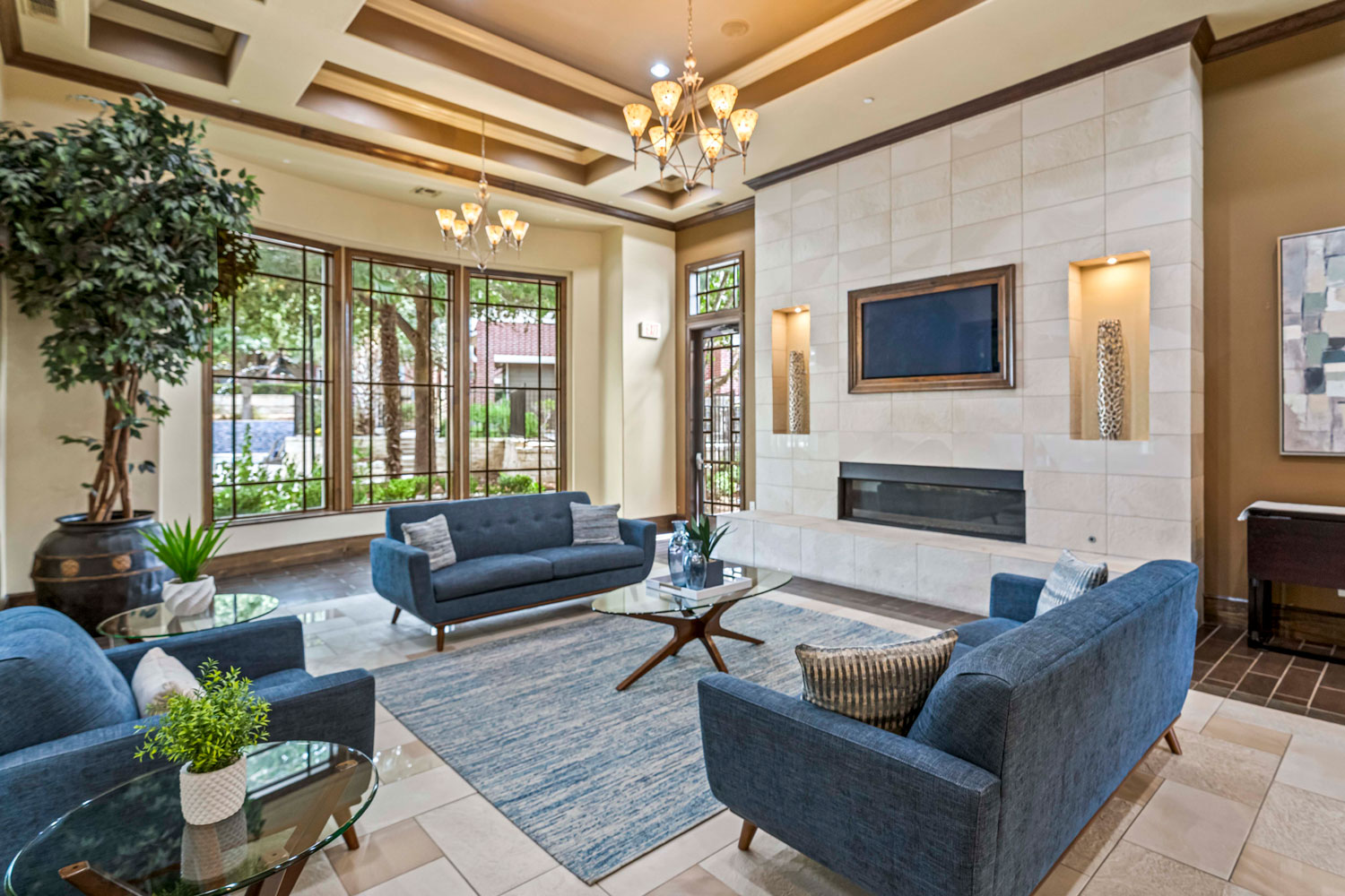 Clubhouse lounge with blue sofas, glass coffee table, tiled fireplace feature wall, and large windows overlooking landscaped courtyard