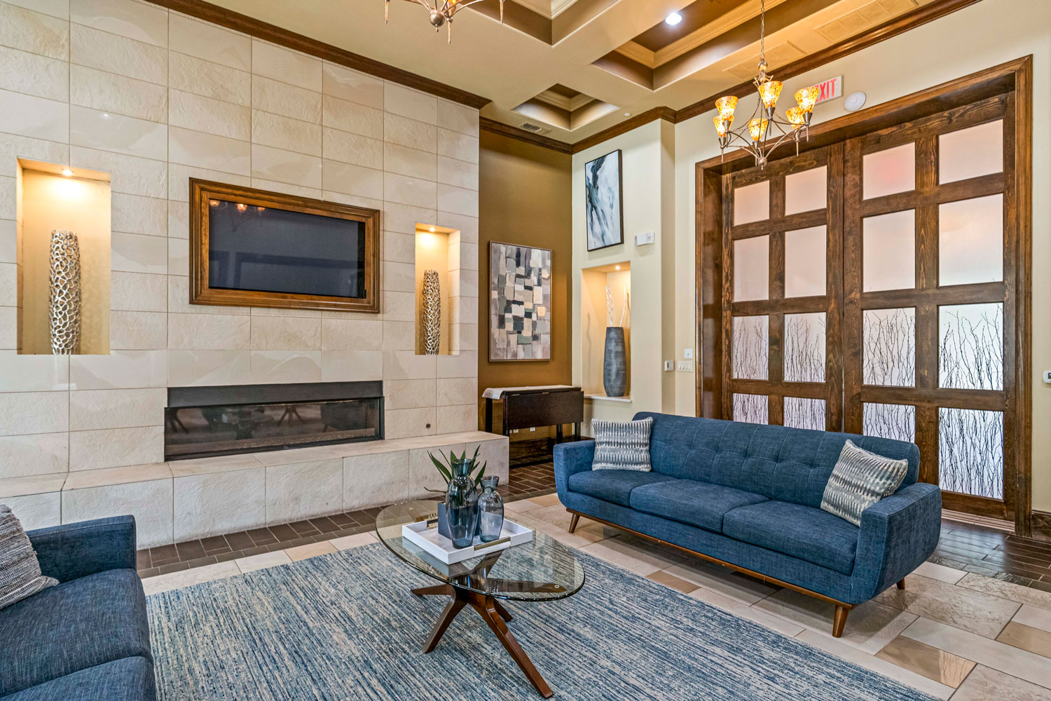 Elegant clubhouse lounge with blue sofas, glass coffee table, tiled fireplace wall, and decorative lighting fixtures